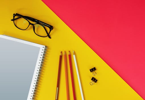 Colorful workspace flat lay with notebook, pencils, glasses, and clips on red and yellow background.